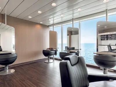 Ein moderner Friseursalon an Bord der Mein Schiff 5 mit Blick auf das offene Meer. Mehrere schwarze Friseurstühle stehen vor großen Fenstern. Die Wände sind hellbraun, der Boden dunkelbraun.