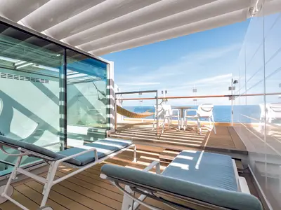 Blick von einer geräumigen Balkonkabine der Mein Schiff 5 mit zwei Liegen im Vordergrund. Im Hintergrund sind eine Hängematte und eine Sitzgruppe auf einem Holzdeck zu sehen, mit weitem Blick auf das blaue Meer unter einem klaren Himmel.