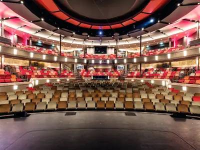 Innenansicht des Theaters auf der Mein Schiff 5. Von der Bühne aus blickt man auf Reihen von roten, orangefarbenen und beigen Sitzen, die in mehreren Ebenen angeordnet sind. Die Beleuchtung ist hell, und das Theater wirkt modern und einladend.