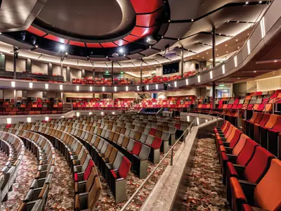 Innenansicht des Theaters auf der Mein Schiff 5, von vorne nach hinten fotografiert. Das Theater ist leer, mit Hunderten von Sitzen in verschiedenen Rottönen, Orange- und Grautönen, die in langen, geschwungenen Reihen angeordnet sind. Es gibt mehrere Ebenen von Sitzgelegenheiten, die sich um eine zentrale Bühne winden. Die Decke ist mit roten und grauen Paneelen sowie zahlreichen Scheinwerfern gestaltet. Der Boden ist mit einem gemusterten Teppich bedeckt.