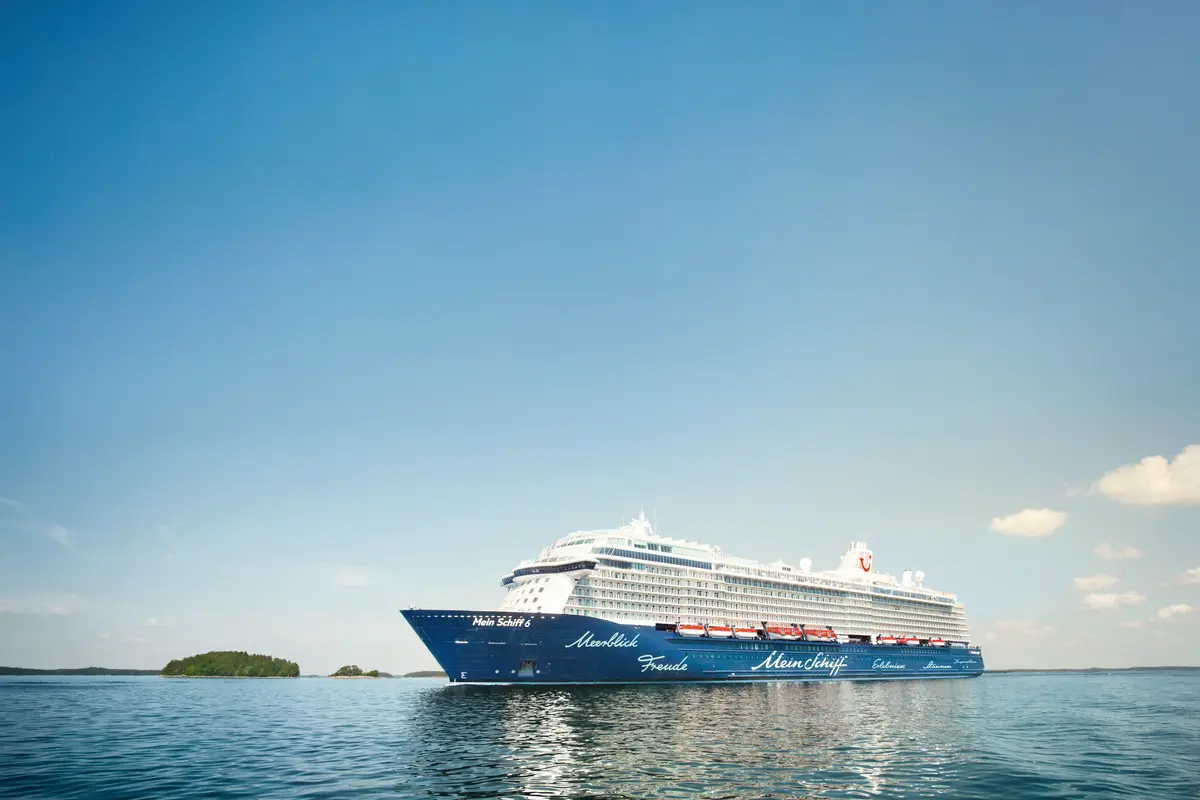 Die blaue und weisse Mein Schiff 6 ist auf ruhigem Meer unter blauem Himmel zu sehen. Grüne Inseln und einige Wolken sind im Hintergrund erkennbar.