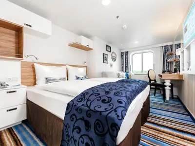 Eine stilvolle Aussenkabine an Bord der Mein Schiff 6 mit blau-gestreiftem Teppich und einem komfortablen Doppelbett. Am Fenster ist ein kleiner Arbeitsbereich eingerichtet.