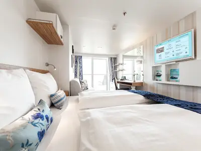 Eine helle und moderne Balkonkabine an Bord der Mein Schiff 6. Zwei Betten sind gemacht, im Hintergrund ein Schreibtisch und ein Balkon. An der Wand hängt ein großer Bildschirm.