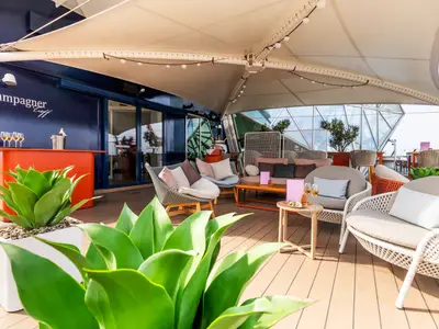 Blick auf den hellen und modernen Champagner Treff an Bord der Mein Schiff 6. Eine überdachte Bar-Lounge mit bequemen Rattanmöbeln, Holzboden und viel Grün.
