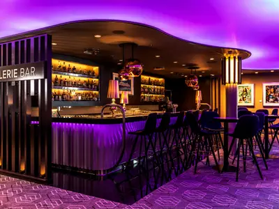 Die moderne Galerie Bar an Bord der Mein Schiff 6. Violett beleuchtete Theke mit Hockern, Spirituosenregalen und Kunstwerken. Elegantes, abendliches Ambiente.