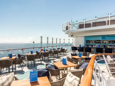 Aussenbar 'Überschaubar' an Bord der Mein Schiff 6 mit Tischen, Stühlen und Barhockern. Meerblick und blauer Himmel prägen die Szene. Die Bar ist bereit für Gäste, ohne Personen.