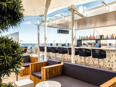 An Bord der Mein Schiff 6: Die Außenalster Bar & Grill mit Meerblick. Blaue Barhocker und bequeme Lounge-Sessel. Die Bar ist bestückt mit Spirituosen, der Himmel ist blau und sonnig.