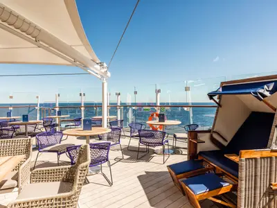 Terrasse der Aussenalster Bar & Grill auf Mein Schiff 6 mit blauen Stühlen, Holztischen und einem Strandkorb. Die Gäste geniessen hier den weiten Meerblick unter blauem Himmel.