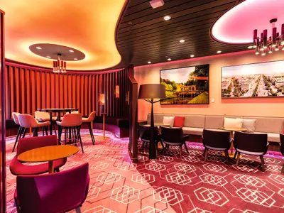 LUMAS Bar an Bord der Mein Schiff 6. Modernes Interieur mit roter Beleuchtung. Loungesessel, Barhocker, Tische, Wandbilder und ein geometrischer Teppich sind zu sehen.