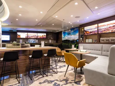 Das helle Café Intención an Bord der Mein Schiff 6. Eine Theke mit Barhockern und Kaffeebohnen-Display. Gemütliche Sitzgruppen mit Tischen und gelben Stühlen. Digitale Menüs und Stadtansichten.