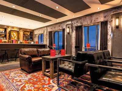 Die gemütliche Studio Bar an Bord der Mein Schiff 6. Braune Ledersofas, schwarze Sessel, eine dunkle Holzbar mit Getränken. Große Fenster und rote Menüs auf Holztischen.