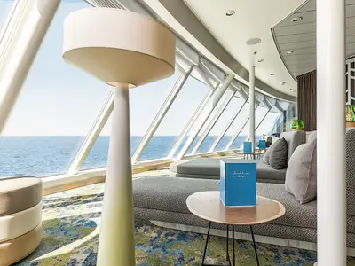 Blick in die moderne Himmel & Meer Lounge an Bord der Mein Schiff 6. Helle Sitzgelegenheiten, Tische und große Fensterfronten bieten einen weiten Ausblick auf das offene Meer unter blauem Himmel.
