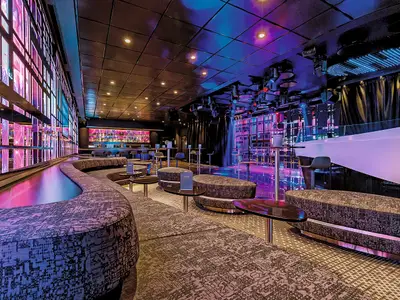 Ansicht der modernen Abtanz Bar an Bord der Mein Schiff 6, beleuchtet mit lila und blauen Lichtern. Gemütliche Lounge-Sitzgruppen und eine Bar sind zu sehen.