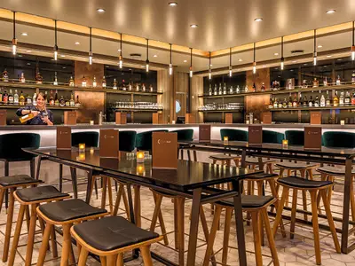 Innenansicht der Schau Bar an Bord der Mein Schiff 6. Eine lächelnde Barkeeperin gießt ein Getränk in ein Glas. Hinter ihr sind Regale voller Spirituosenflaschen zu sehen. Im Vordergrund moderne Bartische und Hocker.