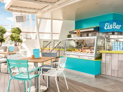 Eis Bar an Bord der Mein Schiff 6. Theke mit Eis und Getränken. Überdachter Außenbereich mit farbigen Stühlen an Holztischen. Pflanzen und helles Tageslicht.