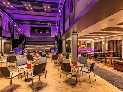 Die Studio Bar der Mein Schiff 6. Ein vielseitiger Raum: links Bühne und gestufte Sitzreihen, violett beleuchtet; rechts Bar-Lounge mit Ledermöbeln und Tischen.