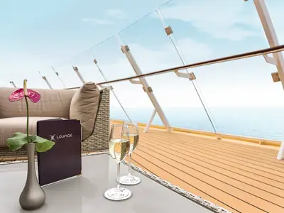 Die sonnige X-Lounge an Bord der Mein Schiff 6. Ein beiges Lounge-Sofa, Couchtisch mit Blume, Speisekarte und zwei Sektgläsern. Meerblick durch eine Glasbalustrade.