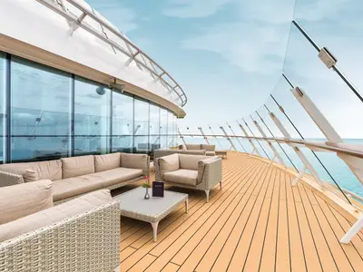 Die Außenterrasse der X-Lounge auf Mein Schiff 6: Helle Rattan-Sitzgruppen mit Polstern bieten Meerblick durch eine Glasbalustrade. Blauer Himmel, sonniger Tag.