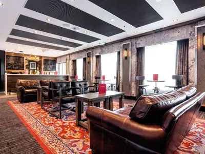 Die Studio Bar an Bord der Mein Schiff 6. Eine moderne und gemütliche Lounge mit bequemen braunen Ledersofas und dunklen Ledersesseln. Holztische stehen auf einem farbenfrohen Teppich. Im Hintergrund befindet sich eine Bar mit Weinregalen und dahinter große Fenster. Das Licht kommt vom Tageslicht und dekorativen Wandlampen.