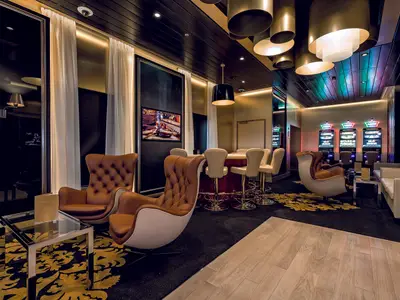 Das Casino und die Lounge an Bord der Mein Schiff 6. Die Szene zeigt Ledersessel, einen Spieltisch und mehrere Slotmaschinen in einem modernen, luxuriösen Interieur.