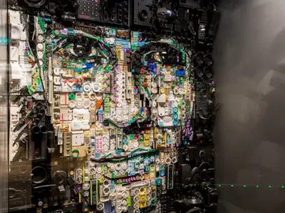 Detailliertes Kunstwerk an Bord der Mein Schiff 6: Ein stilisiertes Gesicht, geschaffen aus unzähligen recycelten Alltagsgegenständen wie Knöpfen, Kabeln und Elektronikteilen. Es ist hinter Glas in einem Innenbereich ausgestellt.
