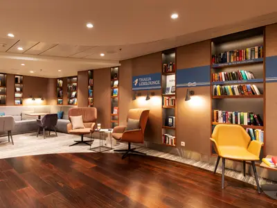 Helle Aufnahme der Thalia Leselounge an Bord der Mein Schiff 6. Bequeme Sessel und Sofas laden zum Verweilen ein, umgeben von beleuchteten Buecherregalen mit vielen Lesestoffen. Die warme Beleuchtung schafft eine gemuetliche Atmosphaere.