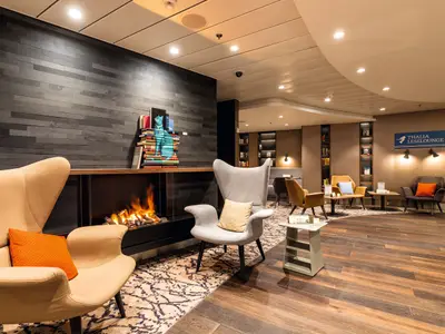 Eine moderne Thalia Leselounge an Bord der Mein Schiff 6 mit Kamin, gemütlichen Sesseln, dunkler Steinwand und vielen Büchern.