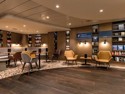 Thalia Leselounge an Bord der Mein Schiff 6. Gemütliche Sessel, Bänke und Tische. Beleuchtete Bücherregale an den Wänden. Dunkler Holzboden und gemusterter Teppichboden.