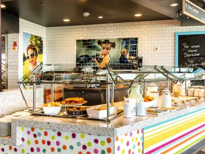 Ein helles, farbenfrohes Kidsbuffet an Bord der Mein Schiff 6. Die Theke bietet Kuchen, Obst und Salate unter Glasschutz. Wandbilder mit lachenden Kindern und bunte Dekoration zieren den Bereich.