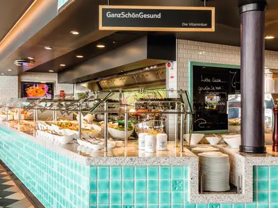 Ein modernes Buffet-Restaurant an Bord der Mein Schiff 6 mit einer langen Auslage voller frischer Speisen. Viele kleine Schüsseln und Servierbesteck sind auf der Theke zu sehen, dahinter ein schwarzes Brett mit Aufschrift und eine offene Küche.