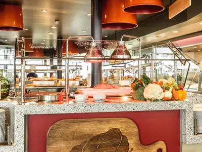 Detailansicht des Anckelmannsplatz Buffet-Restaurants auf der Mein Schiff 6. Zu sehen ist eine Theke mit frischem Fleisch, Gemüse, Saucen und warmen Speisen unter Heizlampen, bereit zur Selbstbedienung.