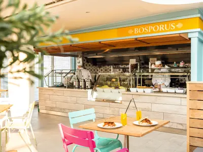 Innenansicht der modernen Bosporus Snackbar an Bord der Mein Schiff 6. Zwei Crew-Mitglieder bereiten Speisen hinter einer Glasvitrine vor, die Salate und andere Gerichte zeigt. Im Vordergrund stehen Tische mit Getränken und Döner-ähnlichen Speisen. Helle, natürliche Beleuchtung durch grosse Fenster im Hintergrund.