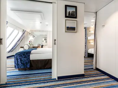 Blick in eine helle Familienkabine an Bord der Mein Schiff 6 mit großem Fenster, einem Doppelbett, Sitzecke und separatem Schlafbereich mit Etagenbetten. Der Teppich hat blaue Streifen.