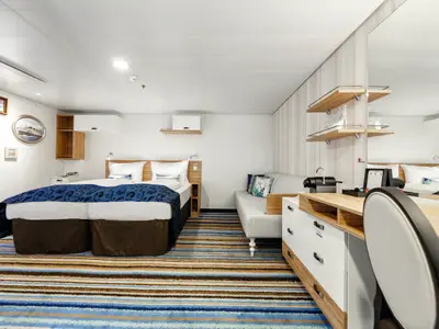 A spacious and modern interior cabin on Mein Schiff 6, showing a double bed, sofa, and desk with coffee machine. The room is decorated with framed pictures and a striped carpet.