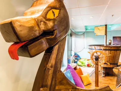 Ein Holzdrachen mit roter Zunge und gelben Augen im Kinderbereich der Mein Schiff 6. Im Hintergrund ein Holz-Spielschiff mit Steuerrad und bunten Kissen.