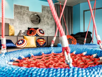 Lebhafter Kinderbereich an Bord der Mein Schiff 6. Großer Netzschaukelkorb und Sofas mit bunten Superhelden-Kissen von Spider-Man, Superman und Captain America.