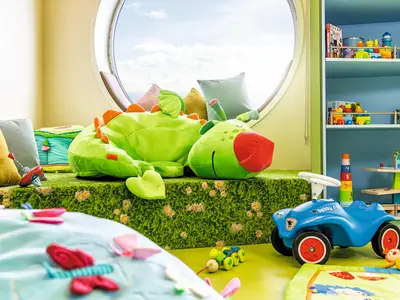Ansicht des Baby-Raums "Nest" an Bord der Mein Schiff 6. Im Raum befinden sich ein grosses gruenes Plueschtier, ein blaues Bobby-Car, Holzspielzeug und ein Bullauge mit Meerblick.