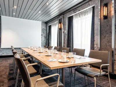 Ein moderner Konferenzraum an Bord der Mein Schiff 6 mit einem langen Holztisch, bequemen Stühlen, einer grossen Projektionsfläche und stilvoller Beleuchtung. Der Raum ist für Meetings vorbereitet.