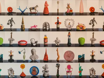 Wandinstallation an Bord der Mein Schiff 6: Eine Vielzahl bunter Kunstobjekte, Skulpturen und Kuriositäten ist symmetrisch auf schwarz-weißen Podesten in einem Raster präsentiert.