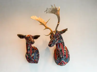 Zwei detaillierte Hirschkopf-Skulpturen, eine mit Geweih und eine ohne, an der Wand an Bord der Mein Schiff 6. Beide sind mit bunten, gemusterten Stoffen bezogen und haben braune Ohren und Schnauzen. Der Hintergrund ist eine helle, ungemusterte Wand.