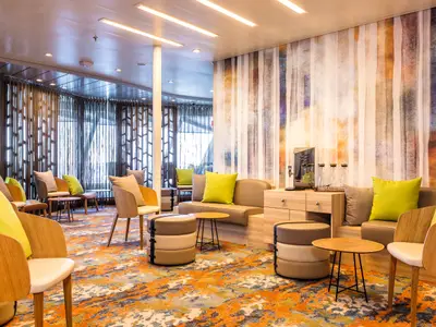 Eine helle, moderne Tee Lounge an Bord der Mein Schiff 6 mit beigen und hellgrünen Kissen auf gemütlichen Holzsesseln und Sofas. Ein farbenfroher Teppich in Orange- und Blautönen bedeckt den Boden. Eine große Fensterfront ist mit dekorativen Gardinen verhängt. An der Wand hängt eine Kunstinstallation in vertikalen Streifen. Eine Tee- und Wasserstation steht bereit.