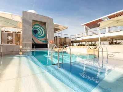 Die Lagunen-Poollandschaft der Mein Schiff 6. Ein großer Pool mit hellem Wasser, Kunstwand mit Mosaik und Uhr. Das sonnige Deck bietet Liegebereiche unter blauem Himmel.