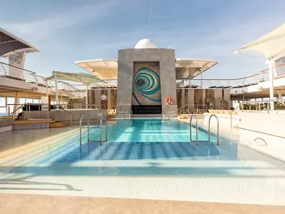 Lagune-Poolbereich der Mein Schiff 6. Großer Pool mit Stufen, flachem Wasser und Spiral-Kunstwerk. Überdachtes Deck. Meer im Hintergrund, keine Personen.
