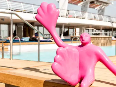 Detailansicht der leuchtend pinken Kunstskulptur Flossi auf dem Holzdeck am Poolbereich an Bord der Mein Schiff 6 bei Tag. Im Hintergrund sind weitere Decks und Badegäste zu sehen.