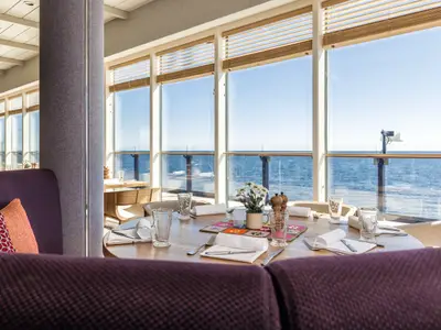 Helles Restaurant an Bord der Mein Schiff 6 mit großen Panoramafenstern und Meerblick. Im Vordergrund ein gedeckter Holztisch mit Blumen und bequemen violetten Sitzen. Die Sonne scheint ins Innere.