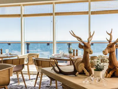 Blick ins Restaurant an Bord der Mein Schiff 6 mit großen Fenstern zum Meer. Zwei Holz-Hirsche und ein Geweih schmücken einen Tisch im Vordergrund. Dahinter weitere Tische mit Stühlen.