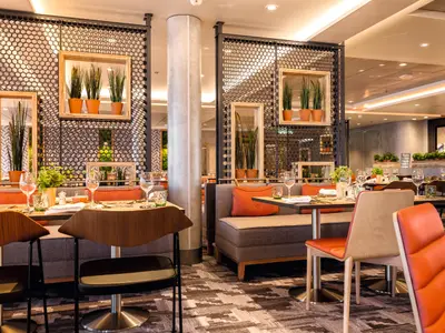 Modernes Restaurant an Bord der Mein Schiff 6. Gedeckte Tische mit orangefarbenen Stühlen und gepolsterten Bänken sind zu sehen. Dunkle Gitterwände mit Pflanzen und Holzrahmen im Hintergrund.