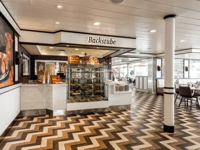 Moderne Backstube auf Mein Schiff 6. Eine Glasvitrine präsentiert Gebäck. Dahinter ein heller Essbereich mit Tischen, Stühlen und Zickzack-Boden.
