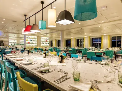 Modernes Restaurant an Bord der Mein Schiff 6. Eine lange, weiss gedeckte Tafel mit Gläsern und Besteck. Bunte Stühle in Türkis und Gelb. Im Hintergrund weitere Tische und Dekorationsteller an der Wand.