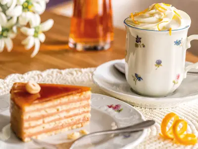 Eine köstliche Cremetorte mit Karamellglasur und ein Getränk mit Sahne und Orangenzesten werden auf elegantem Geschirr an Bord der Mein Schiff 6 serviert. Im Hintergrund weiße Blumen.
